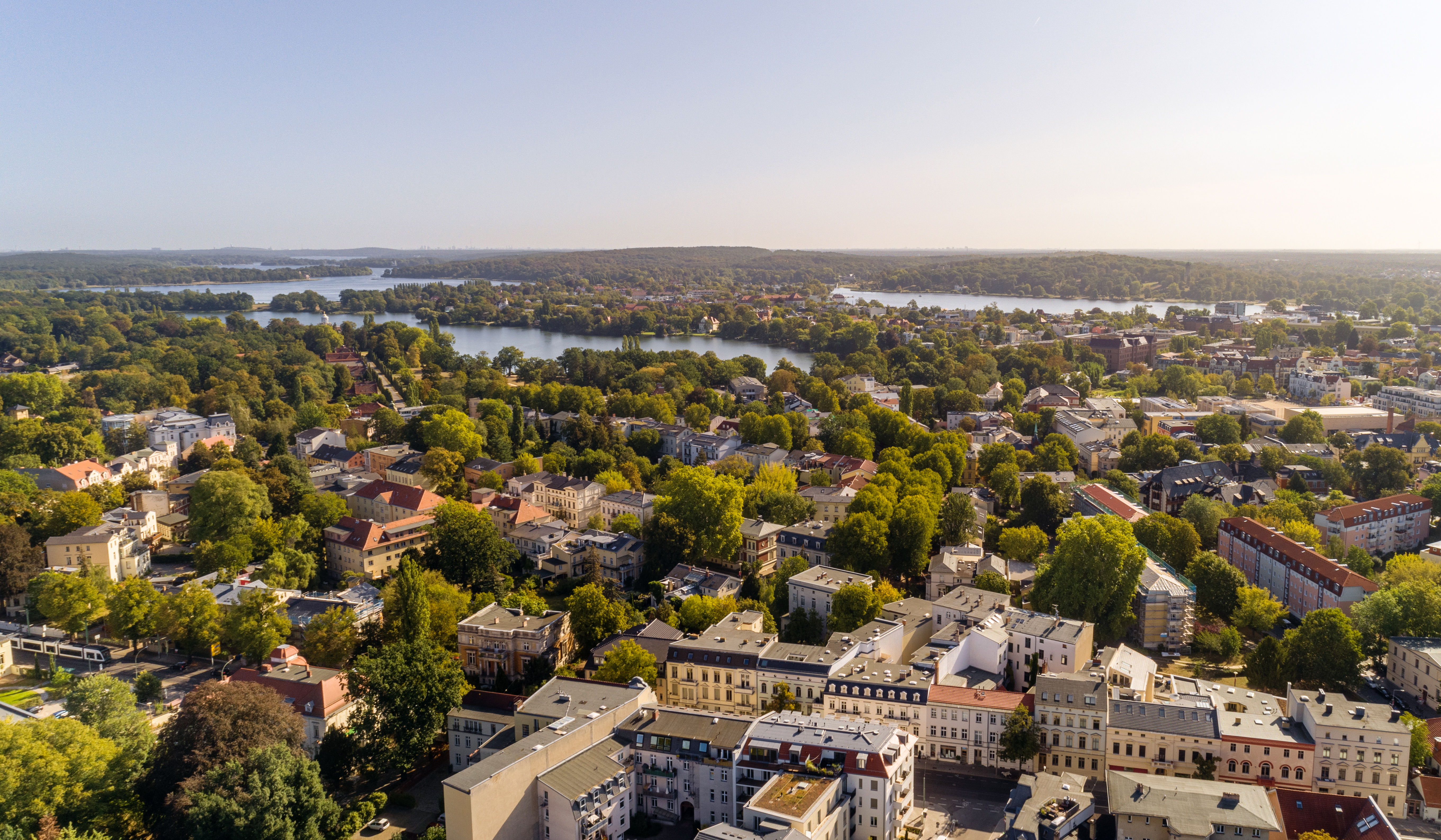 Luftaufnahme einer Stadt mit vielen Bäumen, Gebäuden und mehreren Seen im Hintergrund.