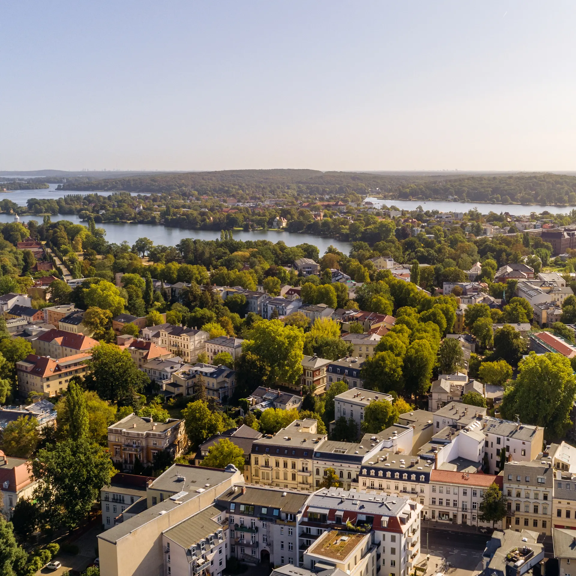 Luftaufnahme einer Stadt mit vielen Bäumen, Gebäuden und mehreren Seen im Hintergrund.