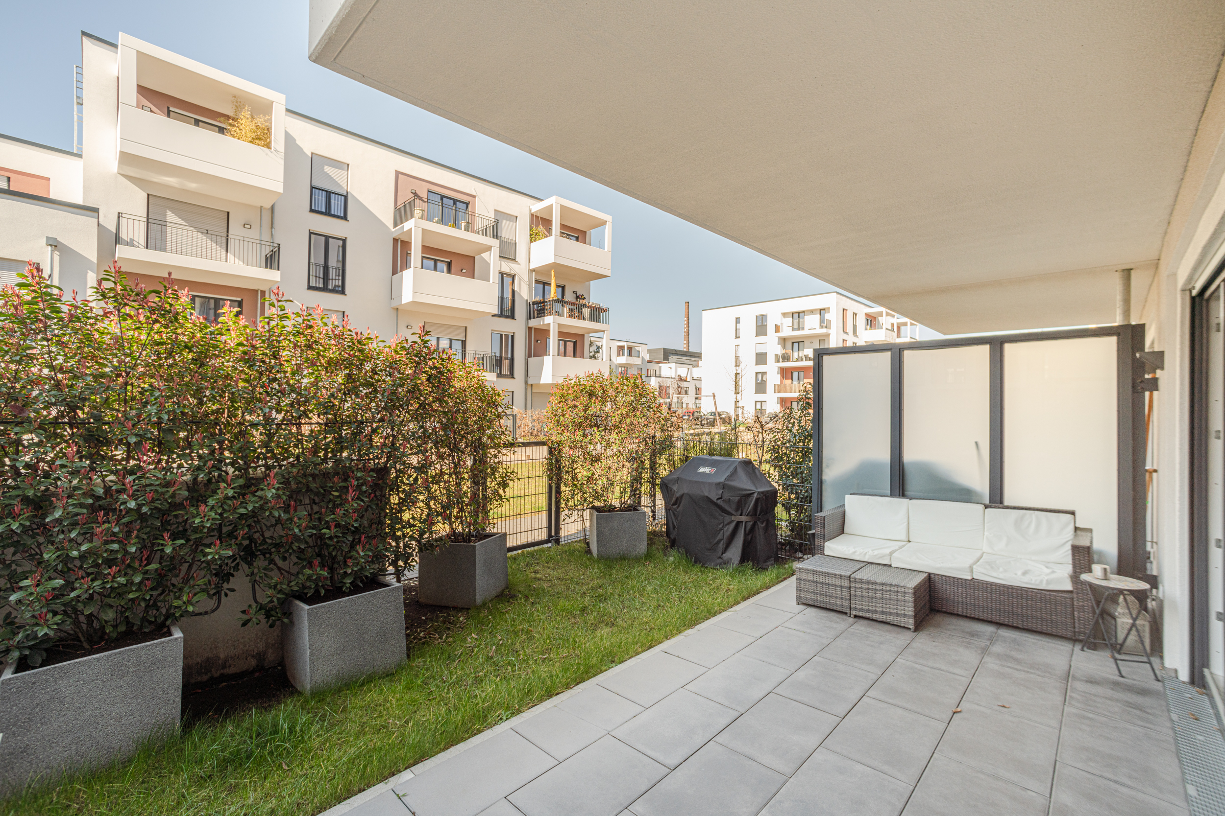 Moderne Terrasse mit Sitzgruppe, Grill und Sichtschutz, umgeben von Hecken und Wohnhäusern.