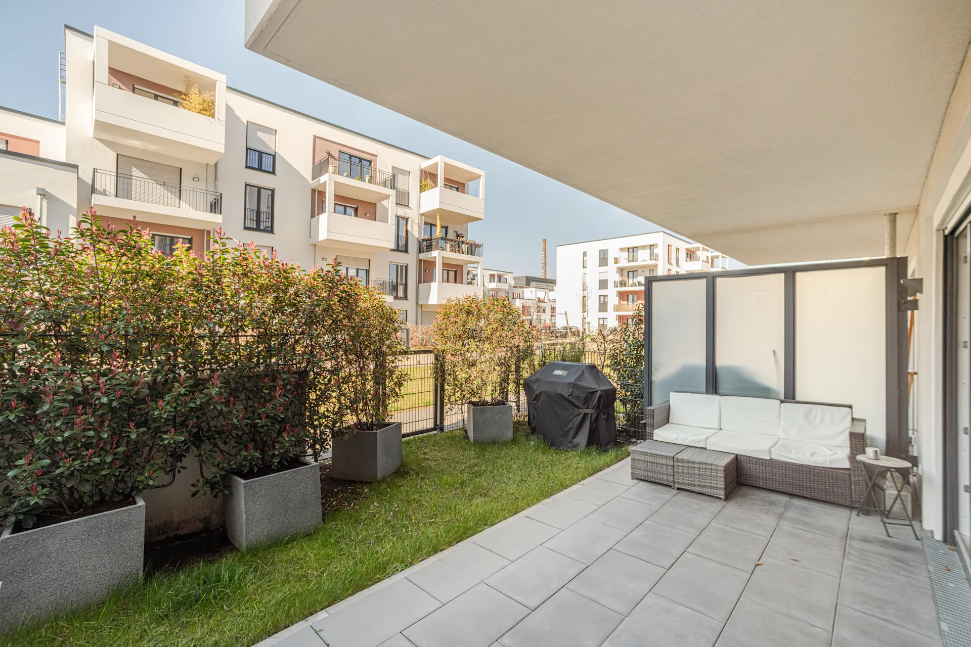 Moderne Terrasse mit Sitzgruppe, Grill und Sichtschutz, umgeben von Hecken und Wohnhäusern.