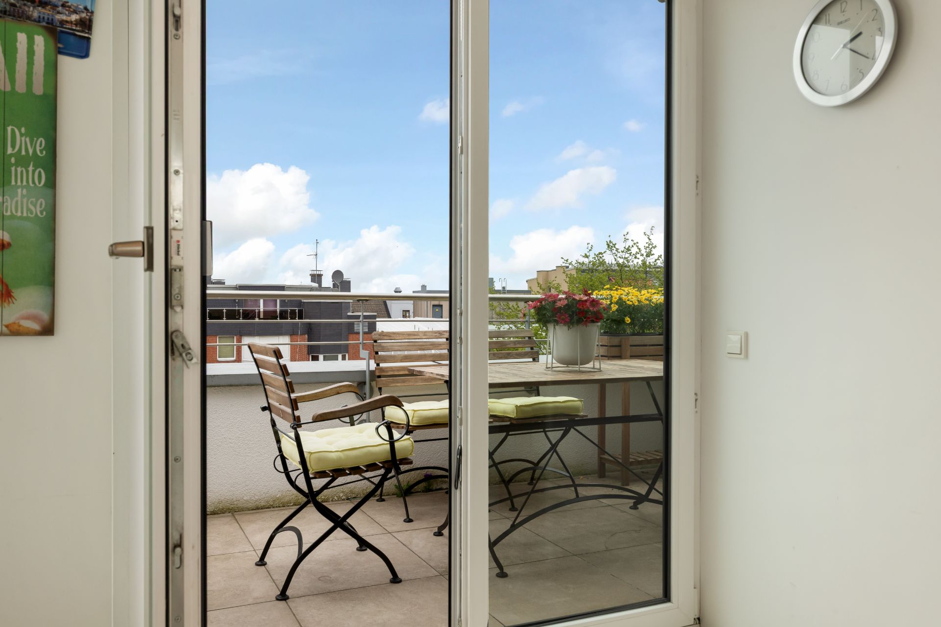 Balkon mit zwei Stühlen, Holztisch und Blumen, gesehen durch eine Glastür.