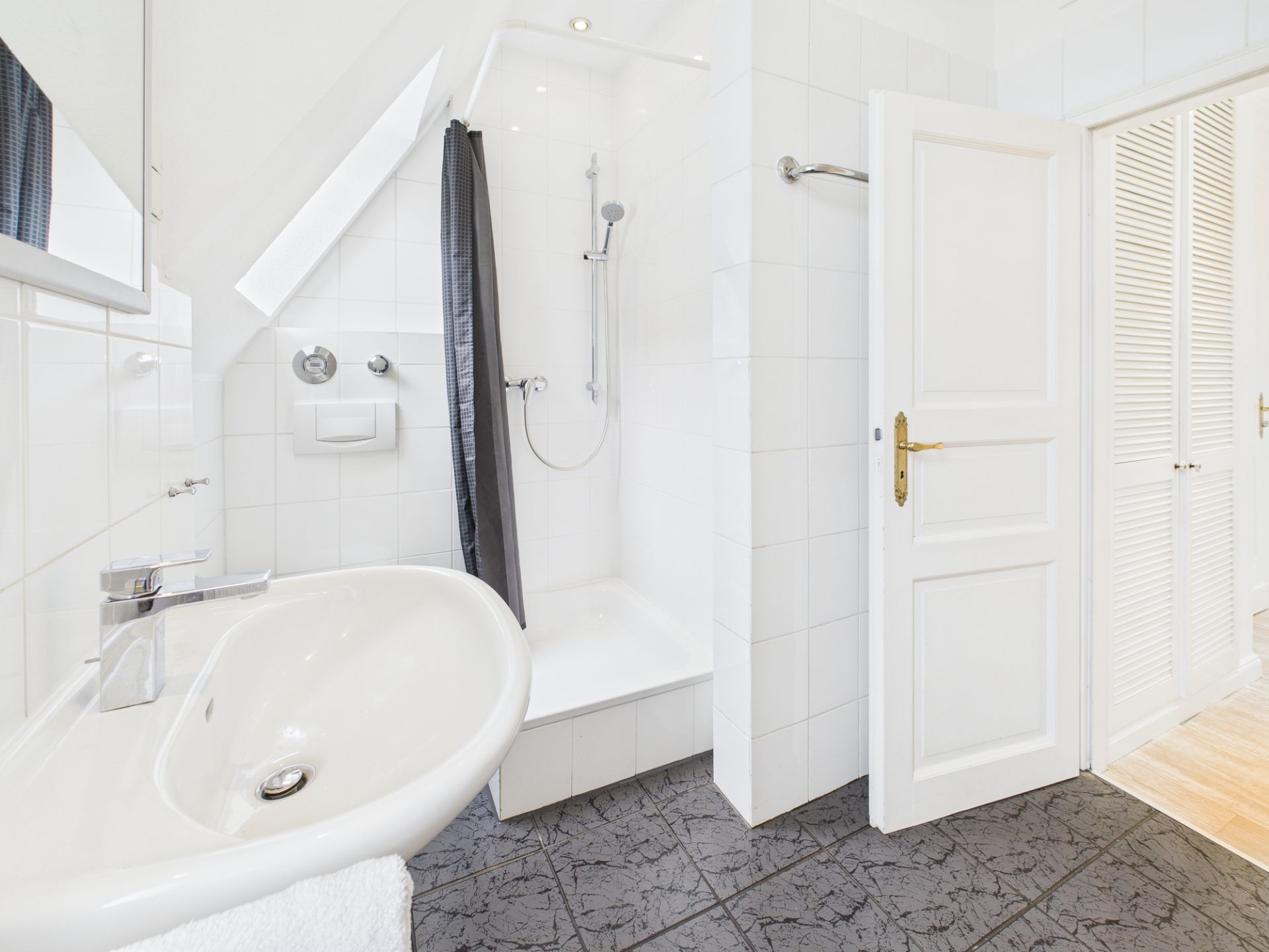 Helles Badezimmer mit Waschbecken, Dusche, offenem Tür und weißen Wandfliesen.