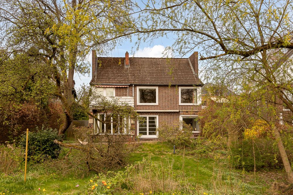 Backsteinwohnhaus mit großen Fenstern, Garten und Bäumen im Vordergrund.