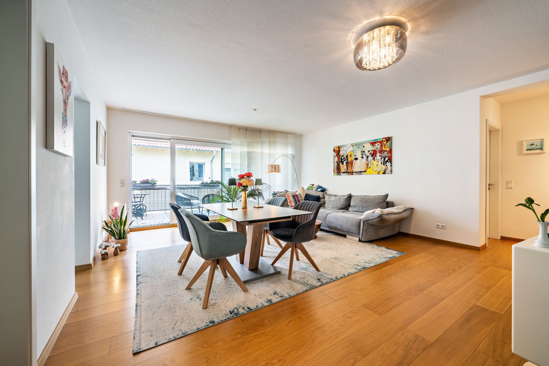 Helles Wohnzimmer mit Esstisch, grauem Sofa, Holzfußboden und Zugang zum Balkon.