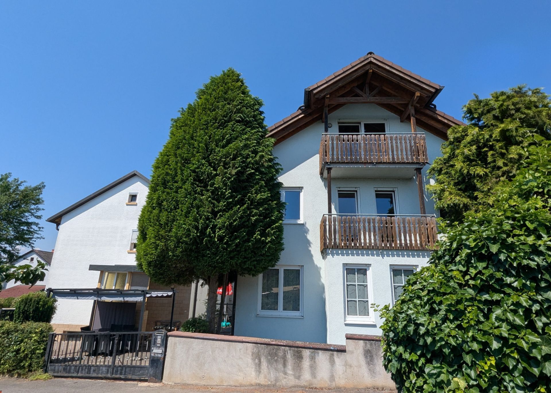 Weiße Wohnhausfassade mit zwei Holzbalkonen und großen grünen Bäumen davor.
