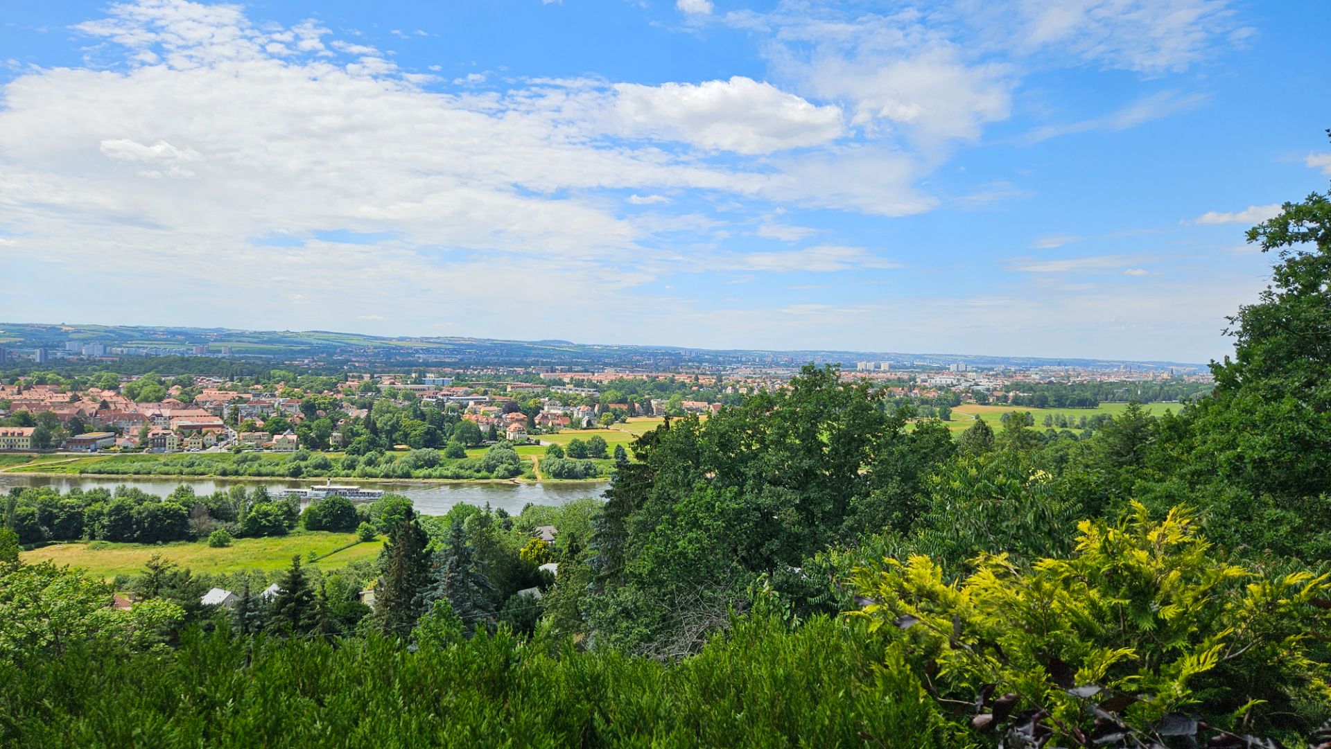 Blick auf Fluss, Bäume und Stadt im Hintergrund unter blauem Himmel mit Wolken.