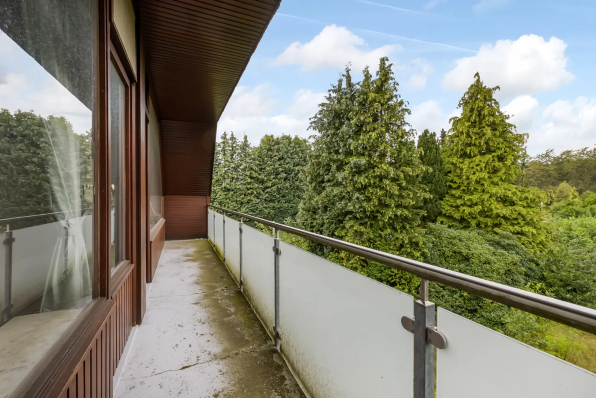 Langer Balkon mit Glasgeländer und Blick auf grüne Bäume.