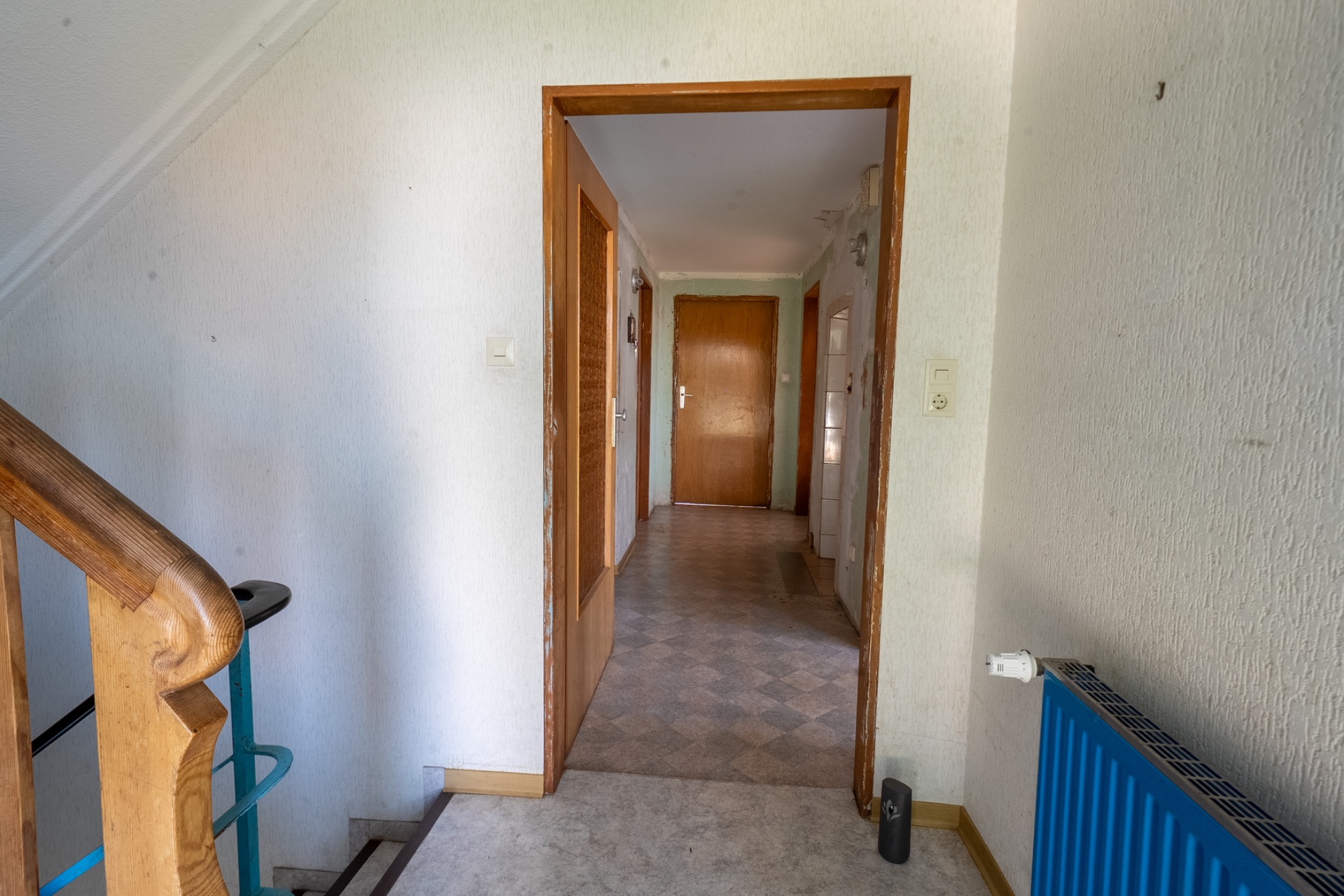 Blick vom Treppenhaus in einen Flur mit mehreren Türen und einem blauen Heizkörper rechts.