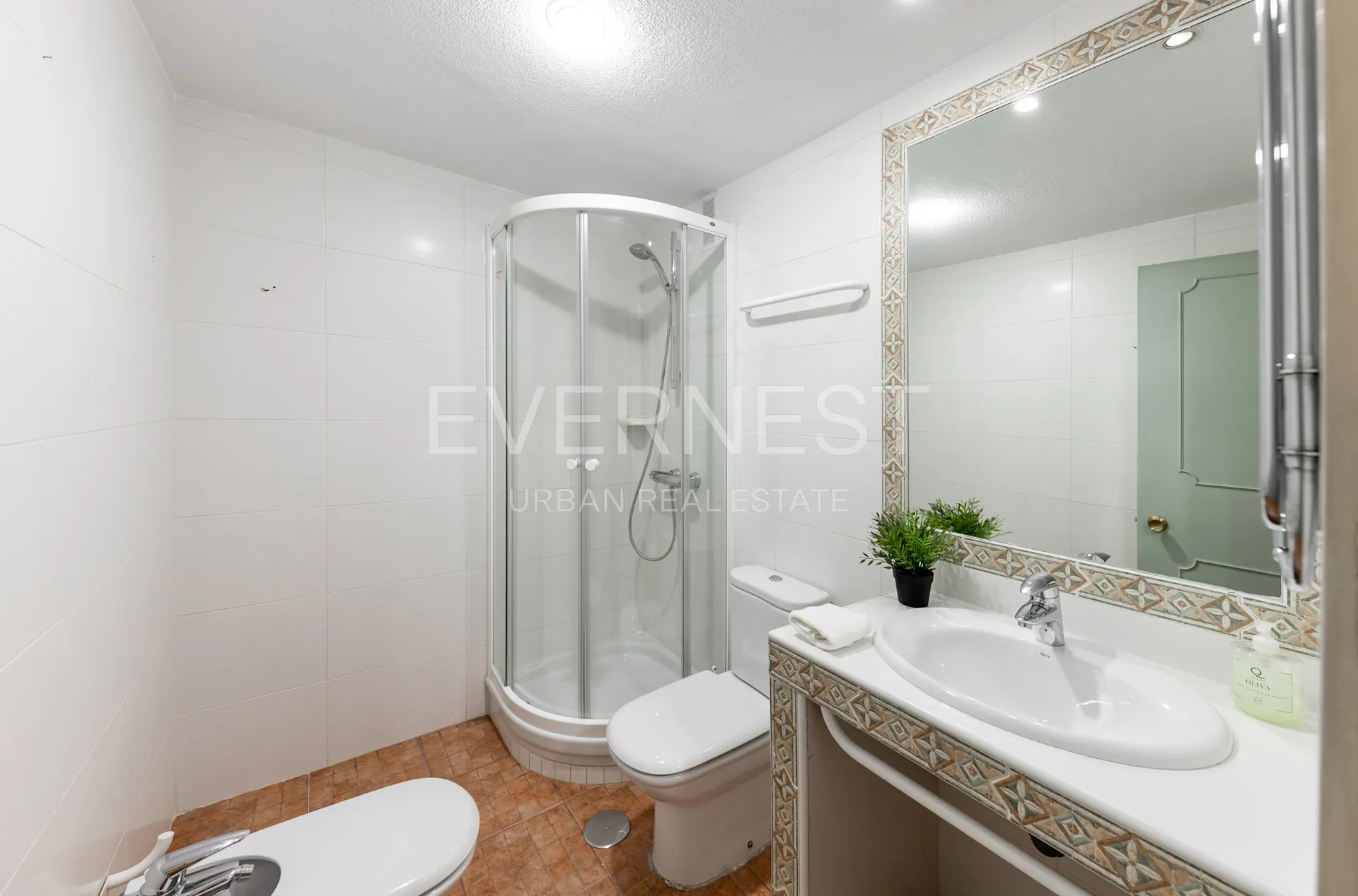 Helles Badezimmer mit Dusche, WC, Waschbecken, großem Spiegel und Pflanze auf der Ablage.