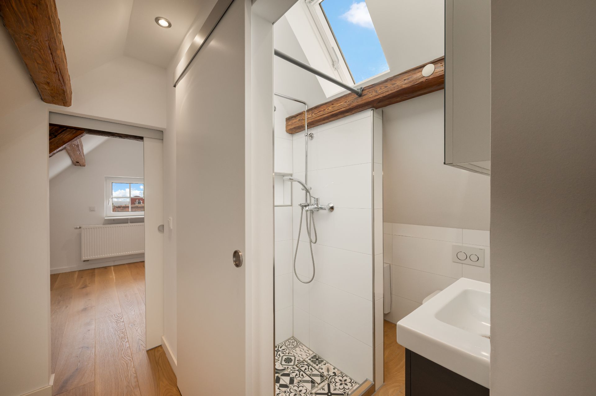 Modernes Badezimmer mit Dusche, Waschbecken und Oberlichtfenster, angrenzend an einen Raum mit Holzboden.