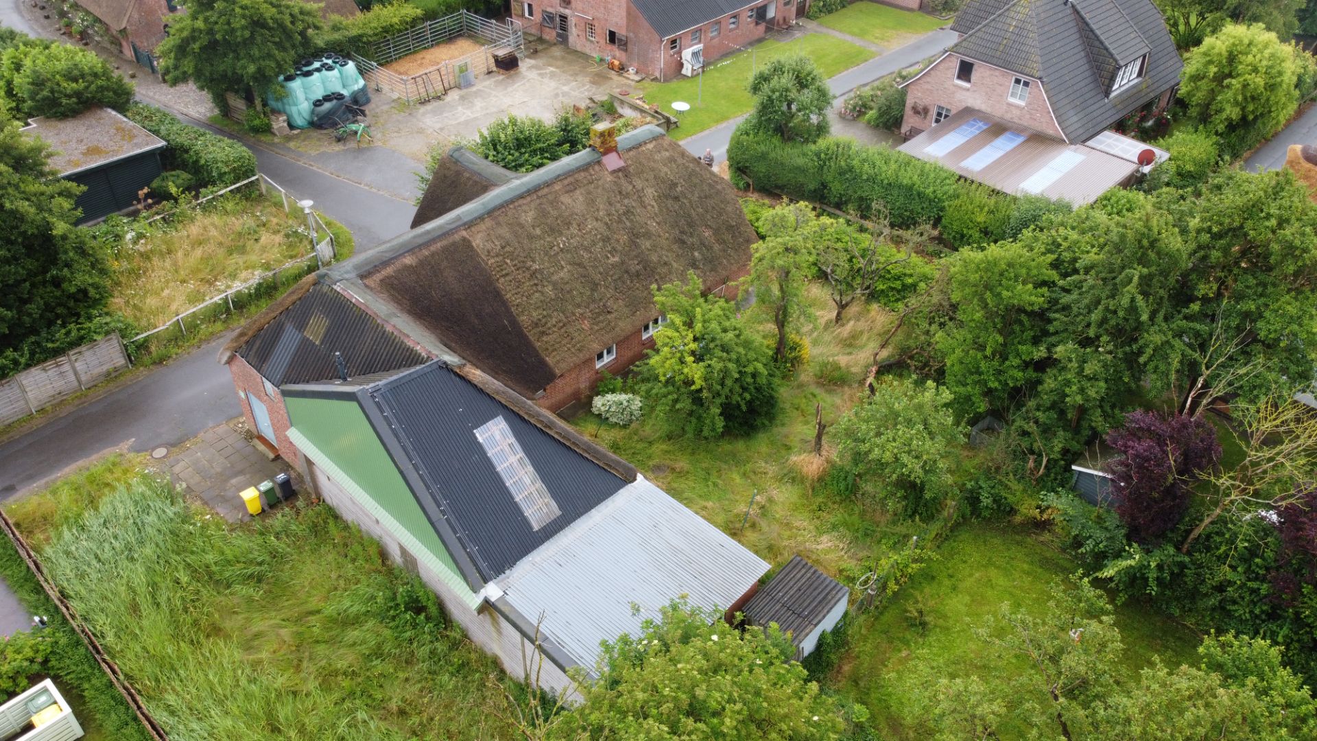 Luftaufnahme eines Hauses mit Reetdach, Nebengebäude und großem Garten am Rand einer Wohnsiedlung.
