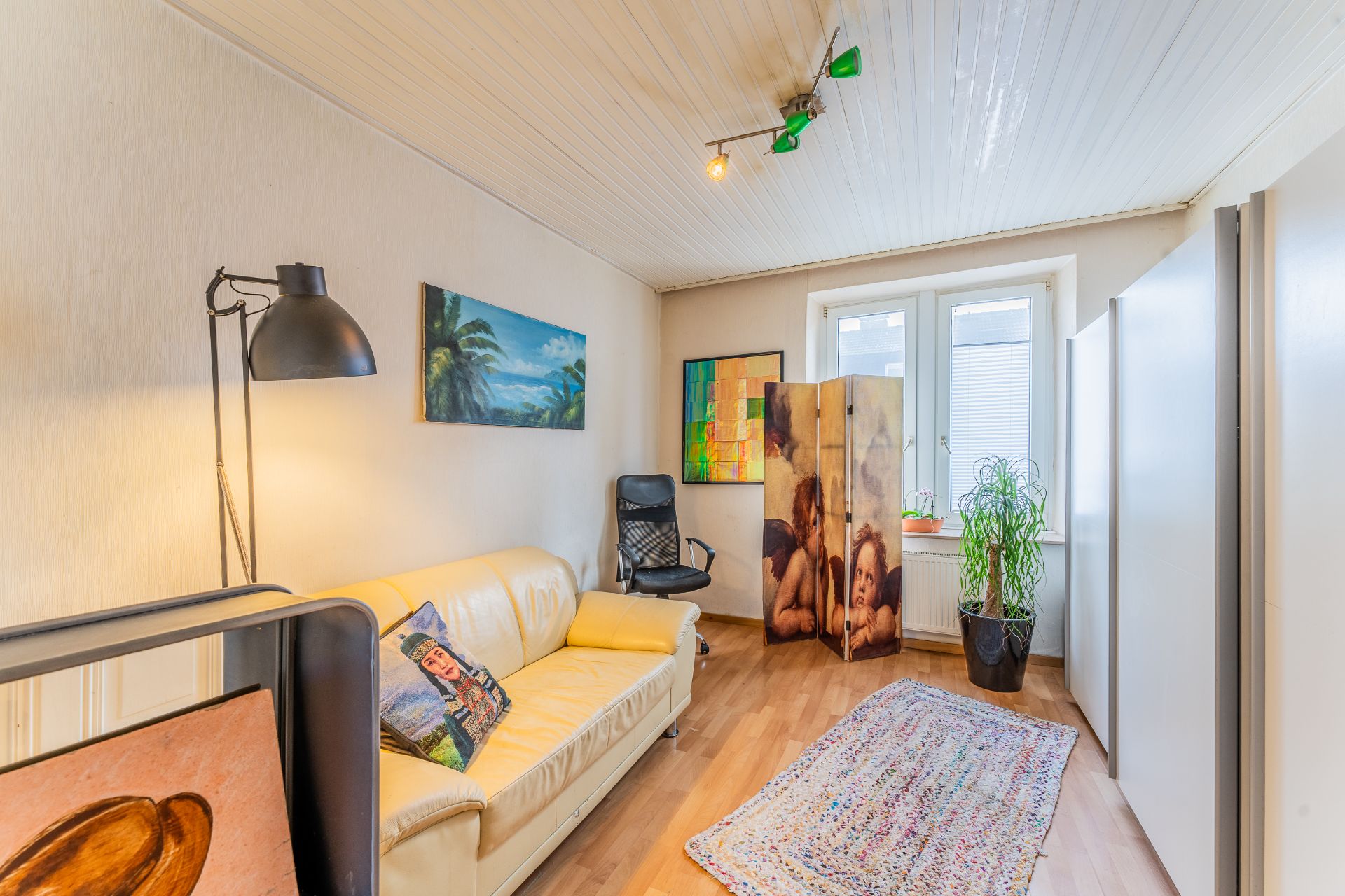 Helles Wohnzimmer mit gelbem Sofa, Stehlampe, Teppich, Pflanze und Paravent vor dem Fenster.