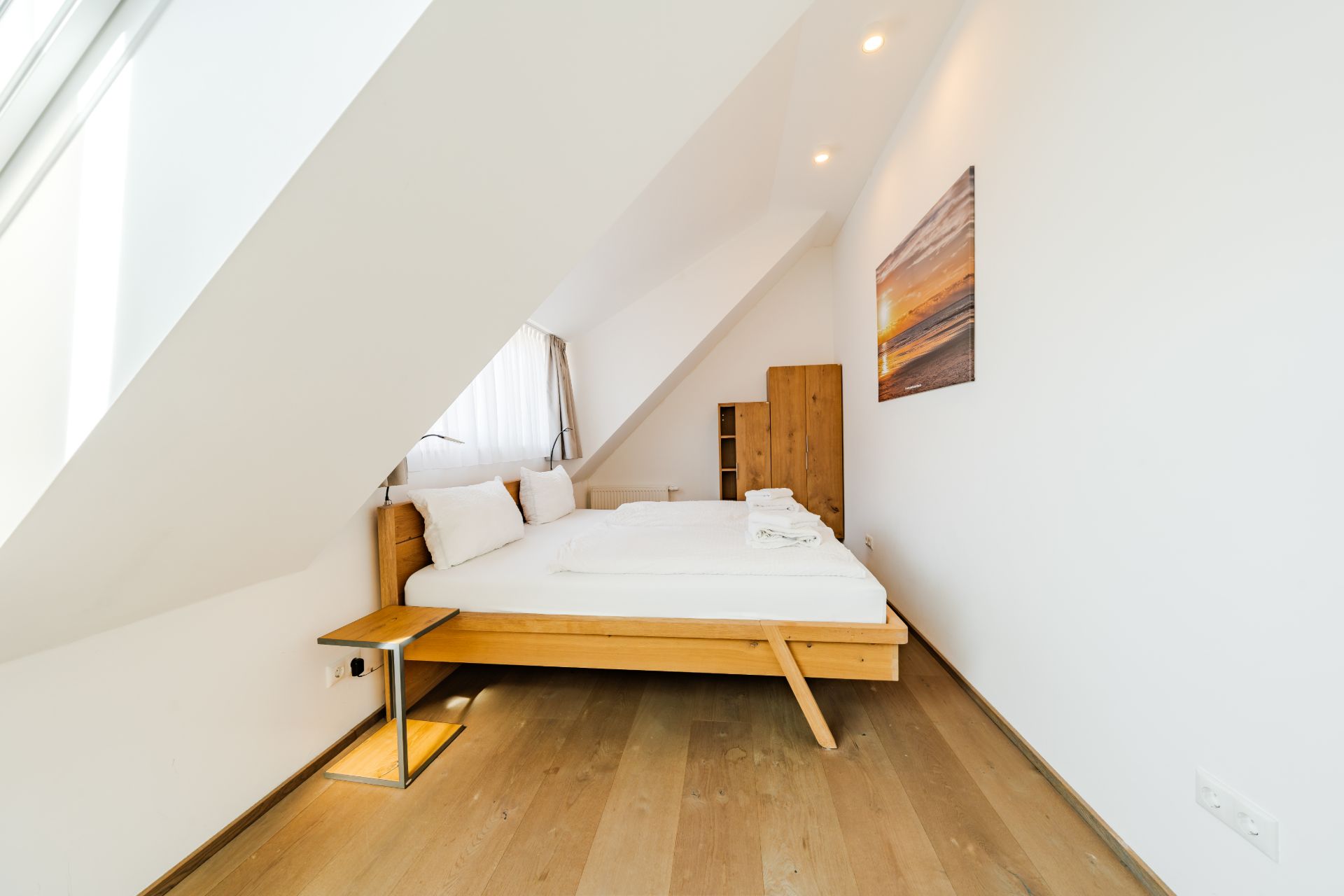 Schlafzimmer mit Holzbett, Nachttisch, Holzschrank und Bild an der Wand unter Dachschräge.