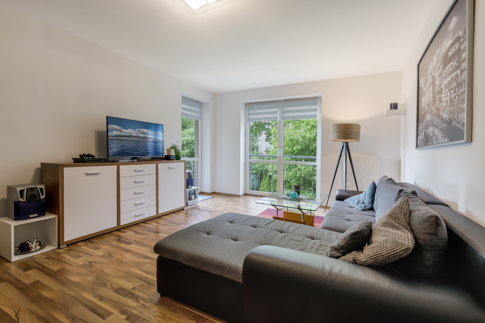 Modernes Wohnzimmer mit grauem Sofa, TV, Sideboard, Stehlampe und großen Fenstern mit Blick ins Grüne.