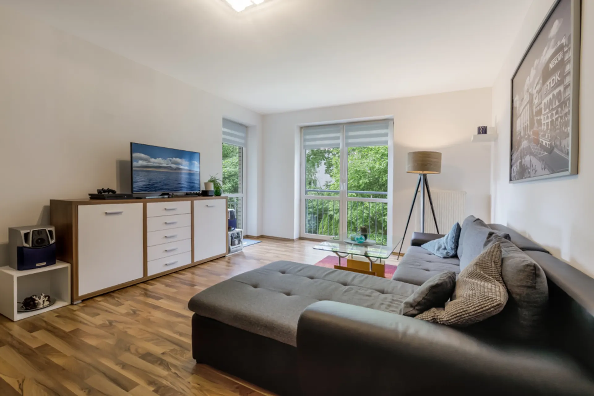 Modernes Wohnzimmer mit grauem Sofa, TV, Sideboard, Stehlampe und großen Fenstern mit Blick ins Grüne.