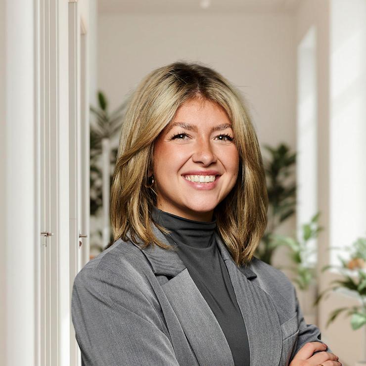 Frau mit blondem Haar in grauem Blazer steht lächelnd in einem hellen Flur mit Pflanzen.