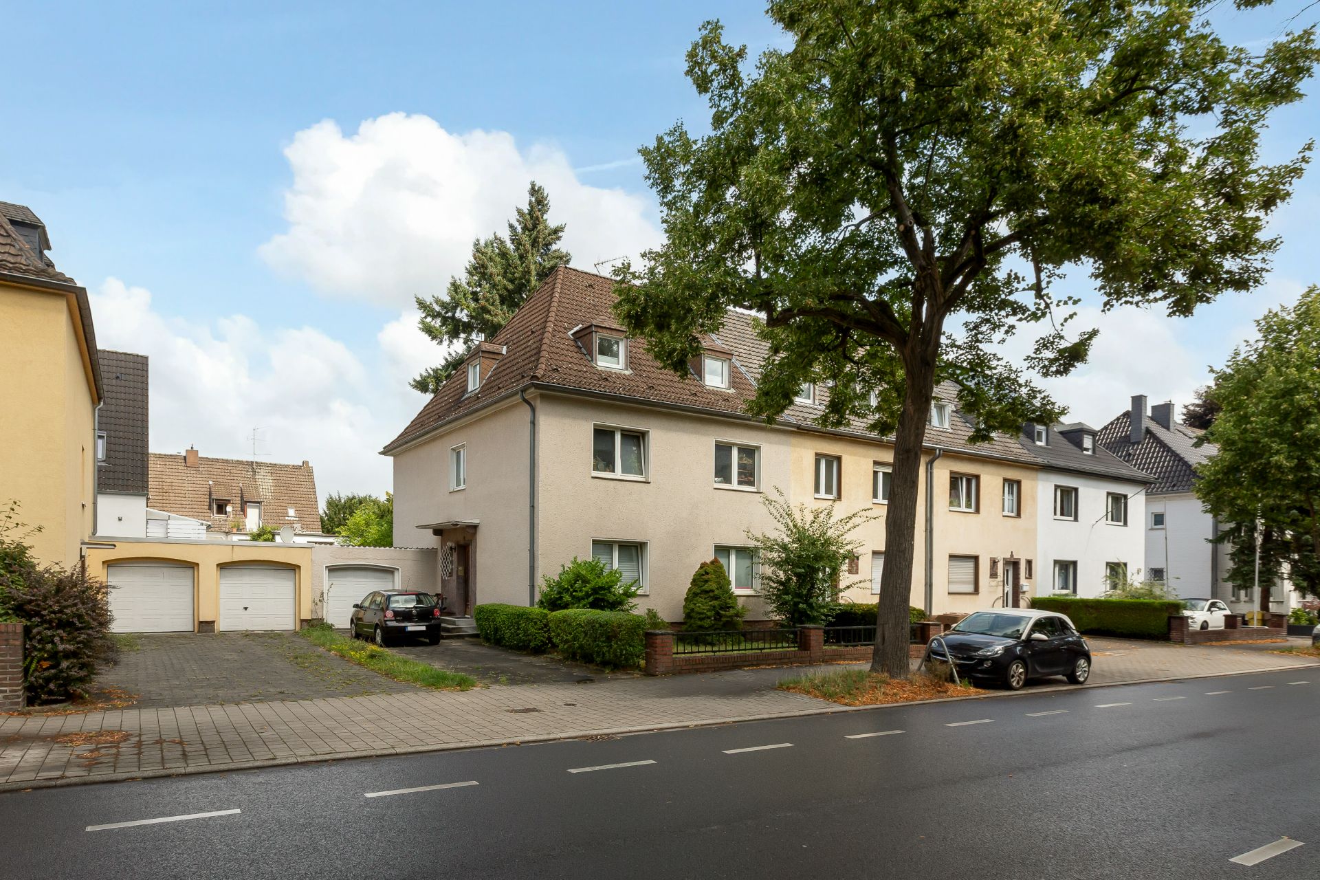 Mehrfamilienhaus mit Vorgarten, Garagen und parkendem Auto an einer ruhigen Straße.