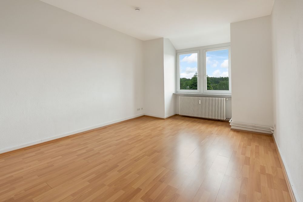 Leerer Raum mit hellem Holzboden, weiß gestrichenen Wänden und großem Fenster mit Blick ins Grüne.