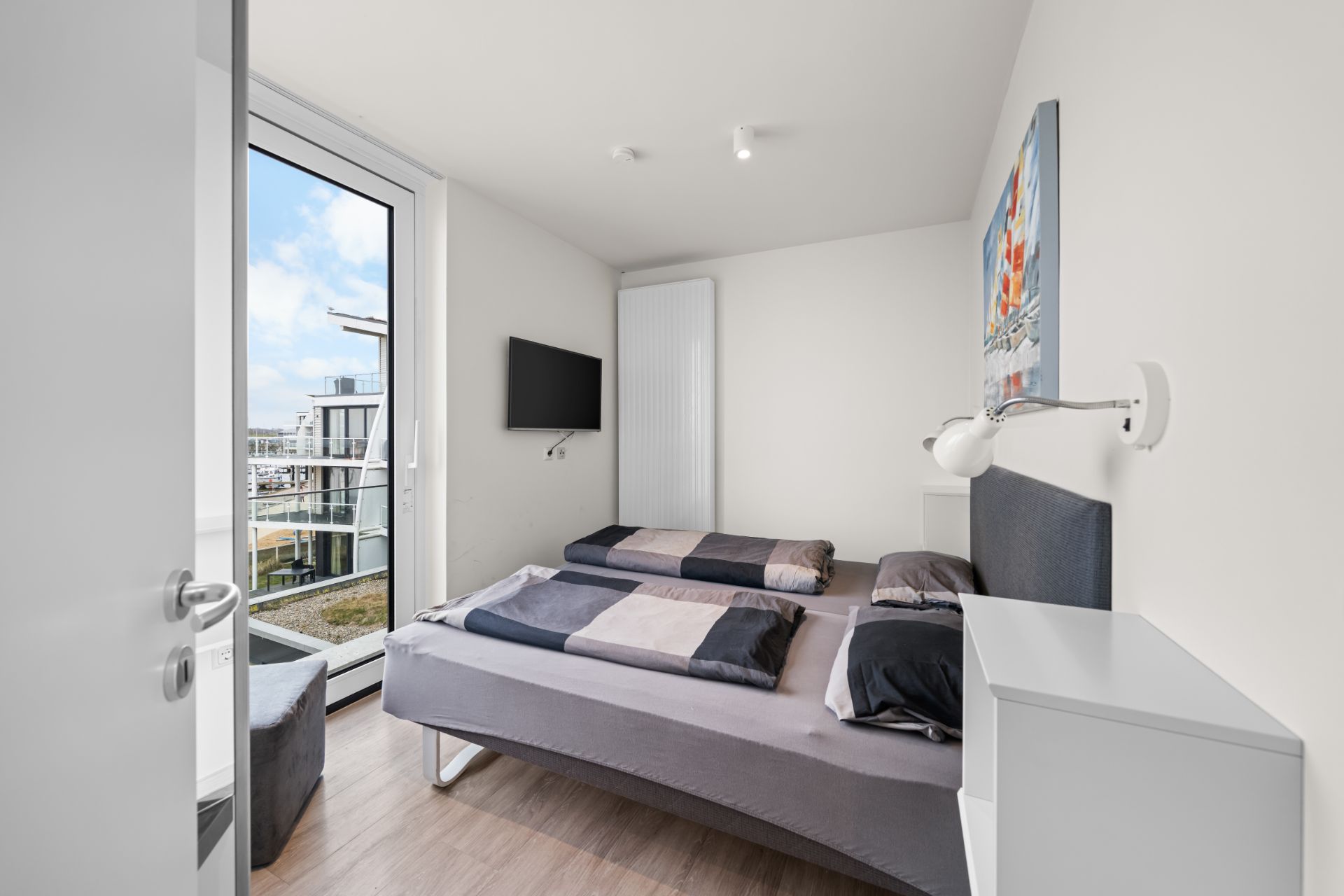 Helles Schlafzimmer mit Doppelbett, Wandbild, Fernseher und Zugang zu einem Balkon.