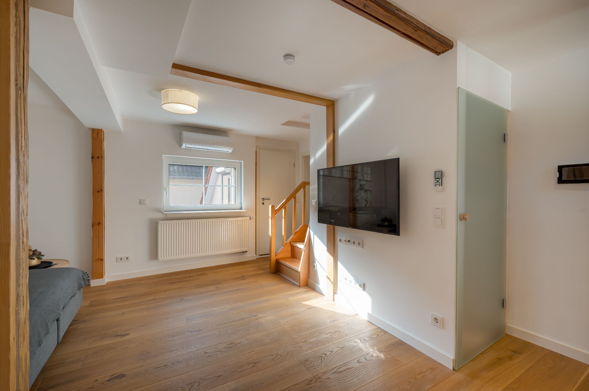 Helles Wohnzimmer mit Holzfußboden, Sofa, Wand-TV und kleiner Treppe neben Fenster.