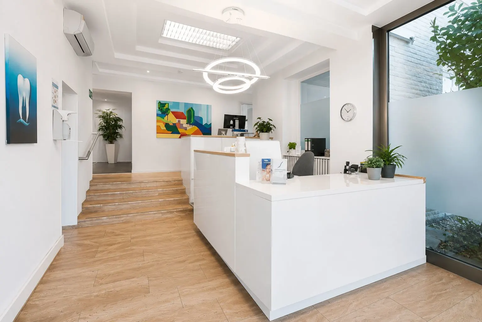 Heller Empfangsbereich mit weißem Tresen, Treppe, Pflanzen und moderner Beleuchtung.