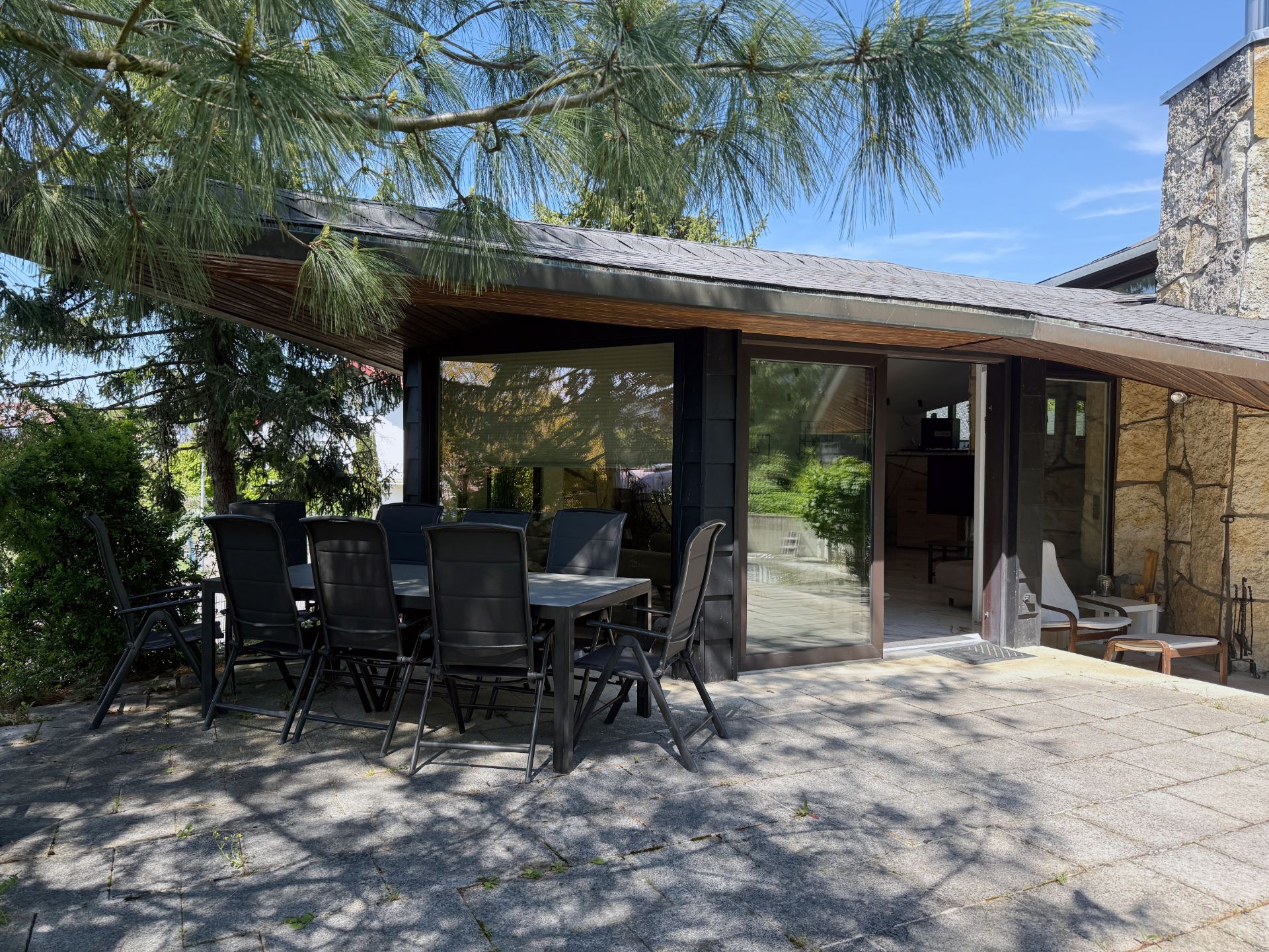 Terrasse mit großem Tisch und acht Stühlen vor einem modernen Wohnhaus, umgeben von Bäumen.