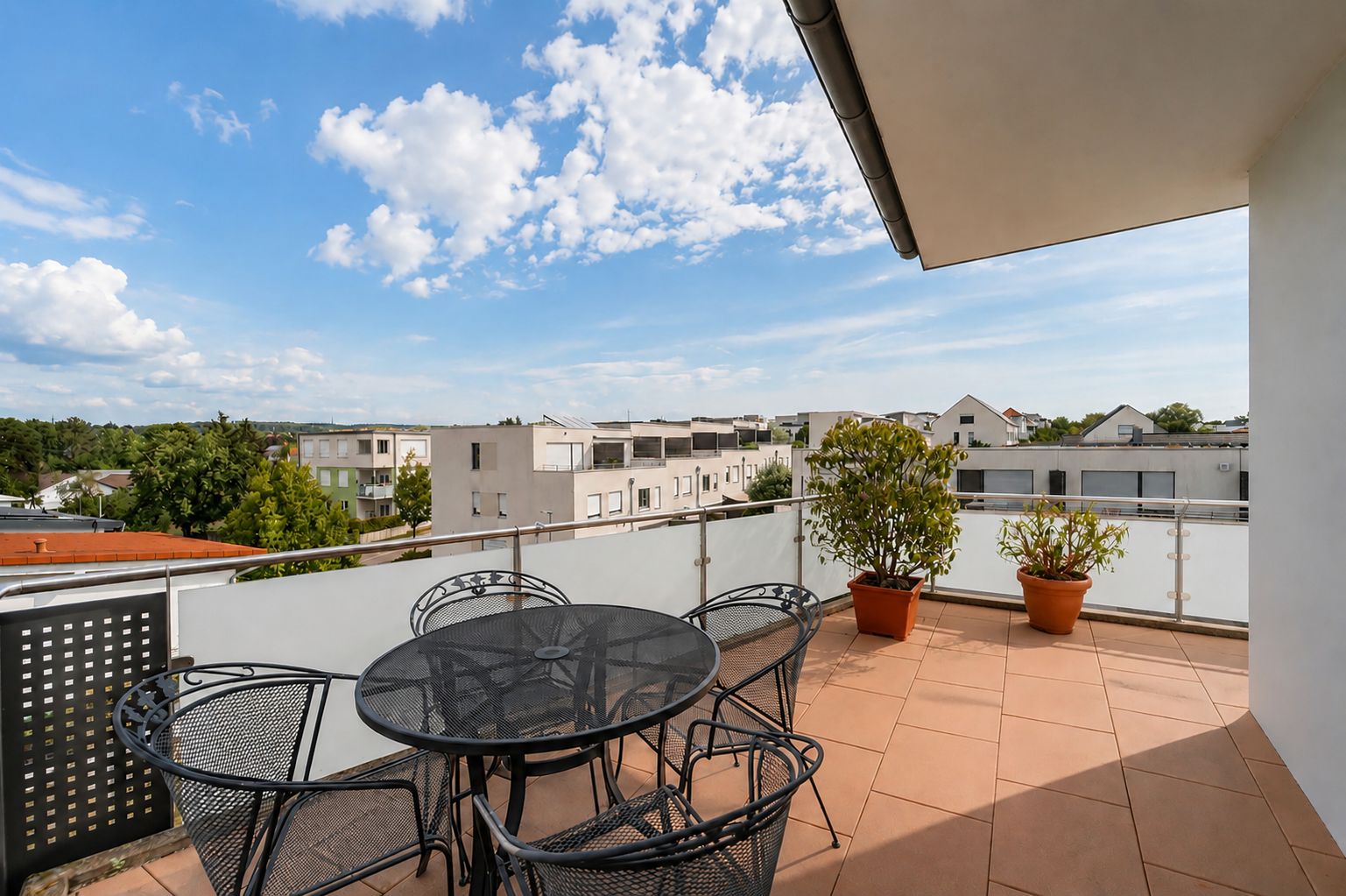 Großer Balkon mit Glastisch, fünf Stühlen und drei Pflanzkübeln, Blick auf Wohnhäuser.