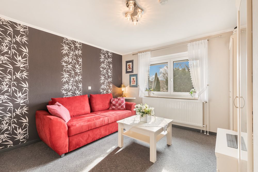 Wohnzimmer mit rotem Sofa, weißem Tisch, gemusterter Tapete und großem Fenster mit weißen Vorhängen.