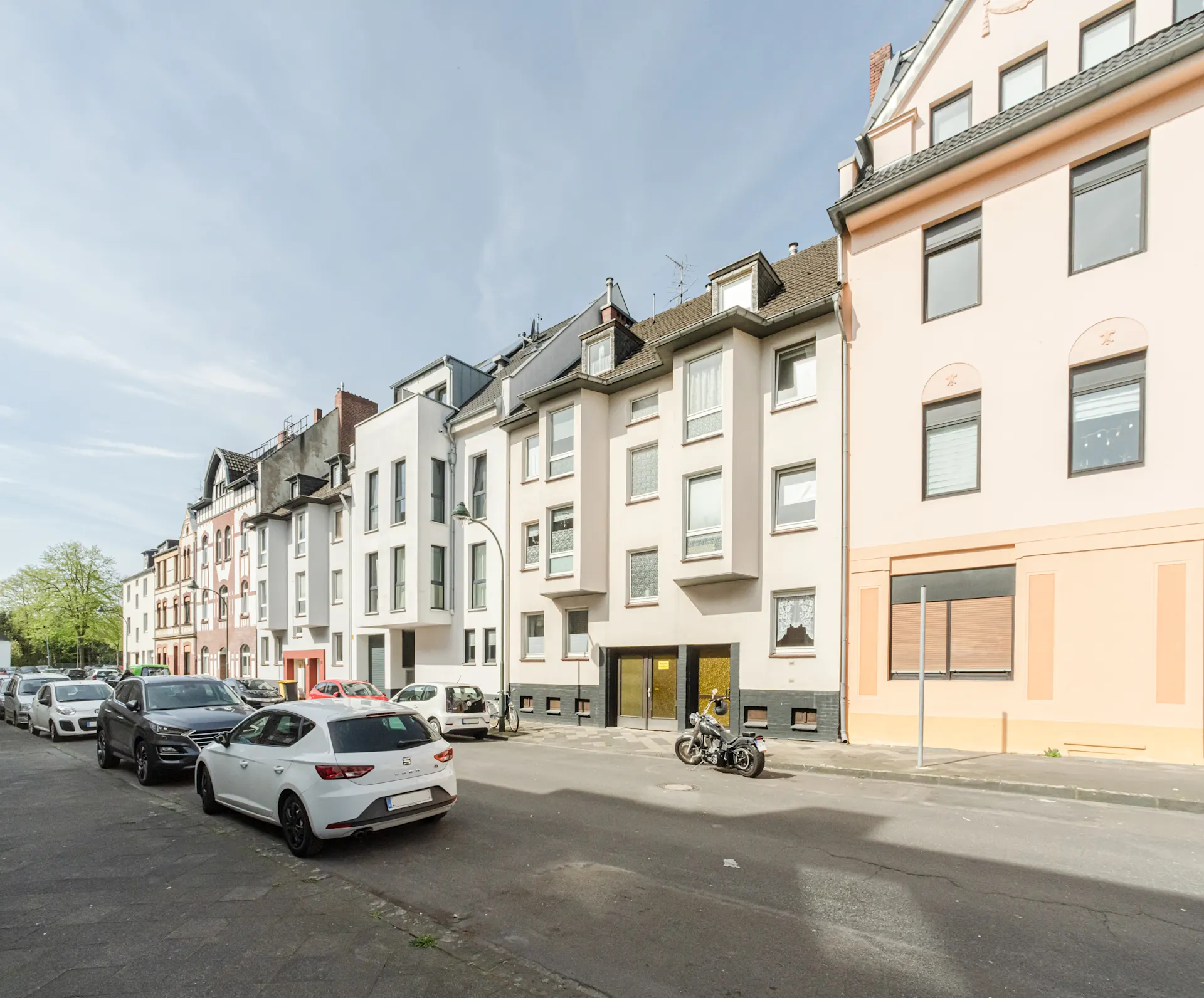 Mehrere Wohnhäuser an einer ruhigen Straße, geparkte Autos und ein Motorrad am Straßenrand.
