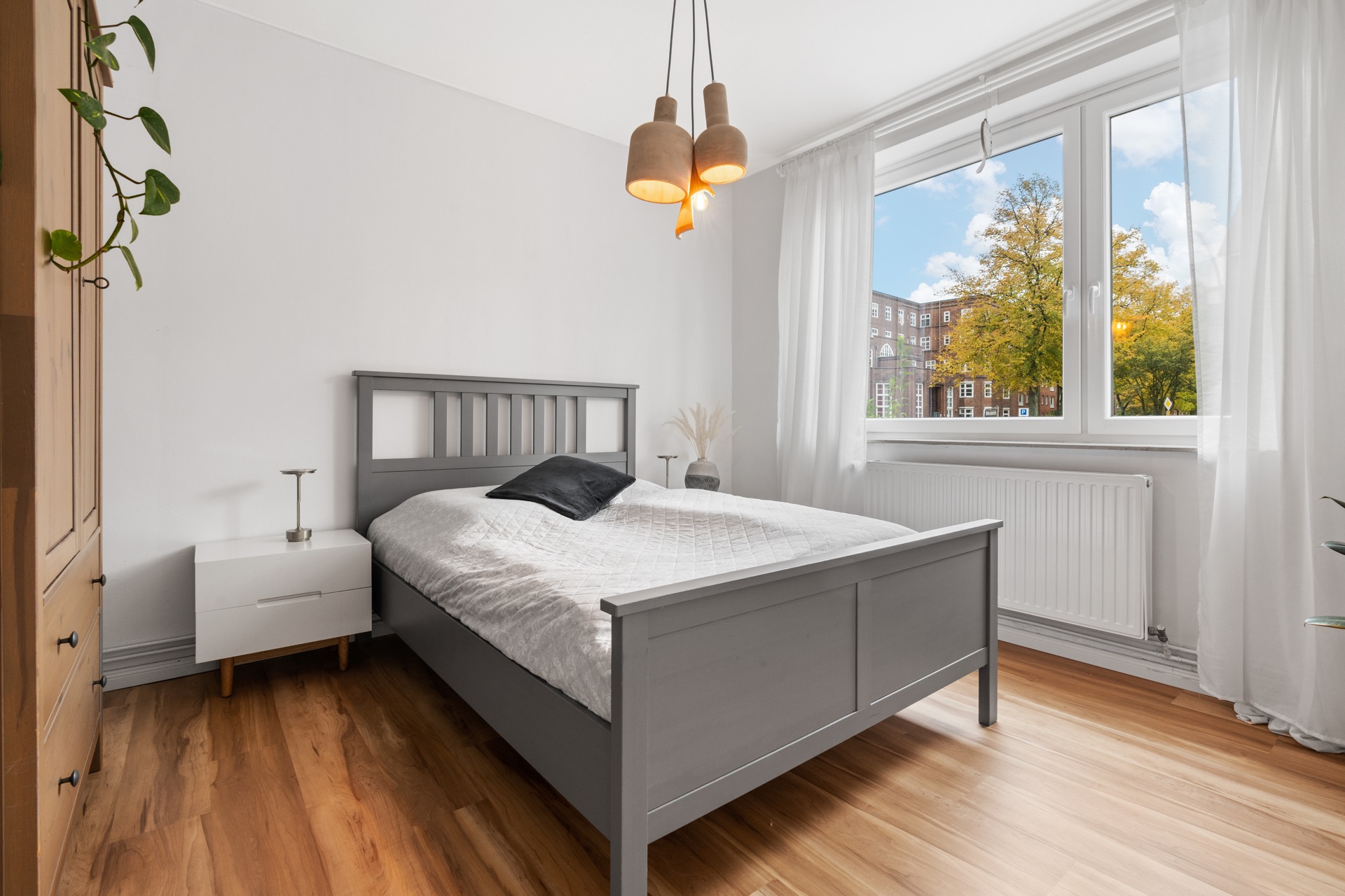 Helles Schlafzimmer mit grauem Bett, Nachttisch, Holzfußboden und großem Fenster.