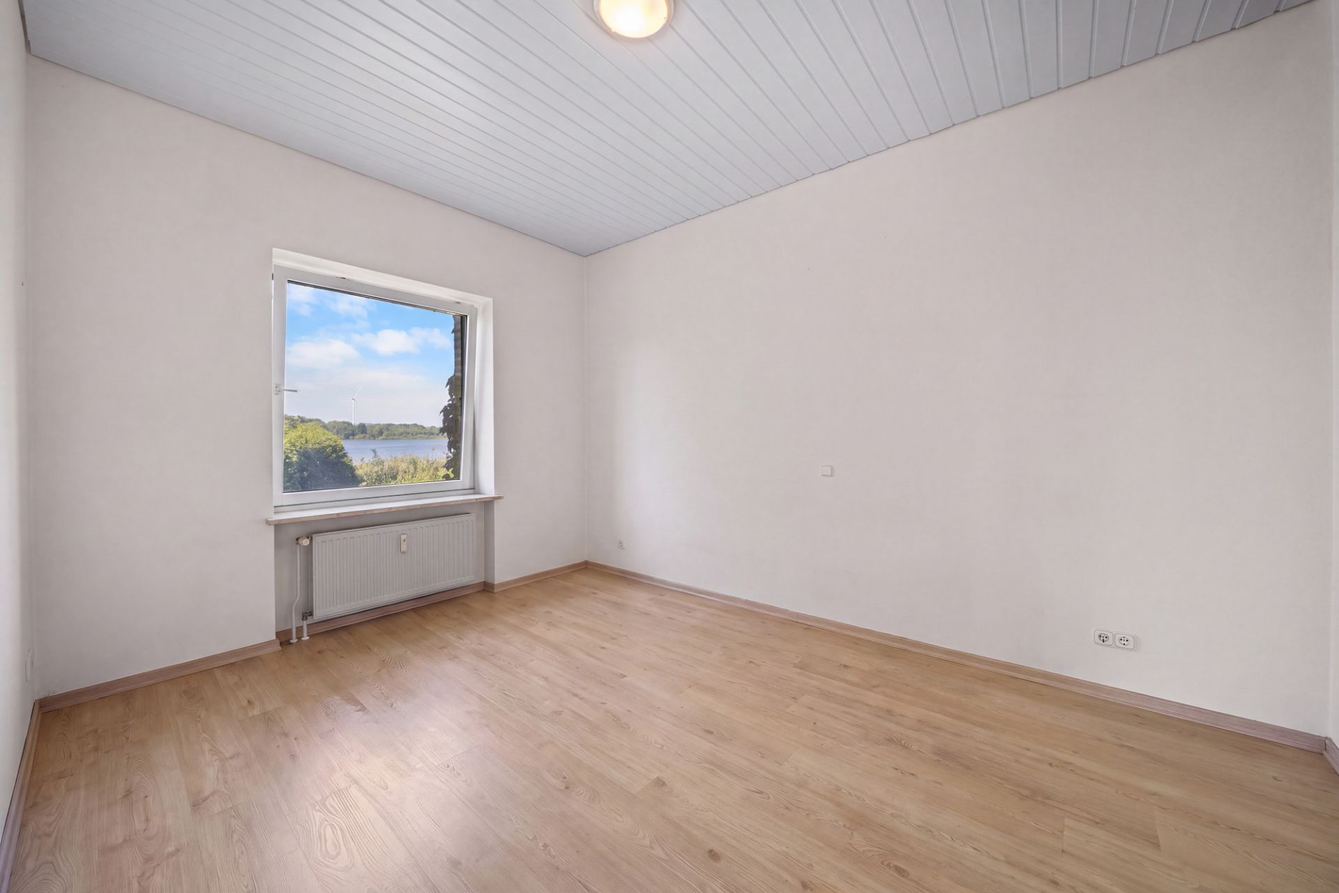 Leerer Raum mit Holzfußboden, weißer Decke und großem Fenster mit Blick ins Grüne.