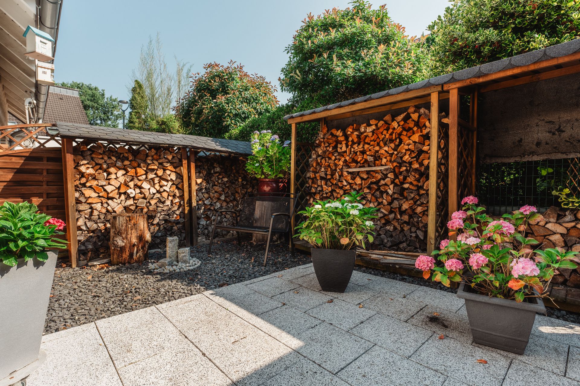 Gepflasterte Terrasse mit Holzlager, Blumen in Töpfen und Bank im Garten.