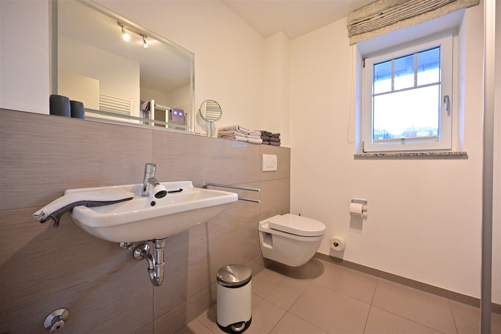 Modernes Badezimmer mit Wandwaschbecken, wandhängender Toilette, Fenster und Spiegel.