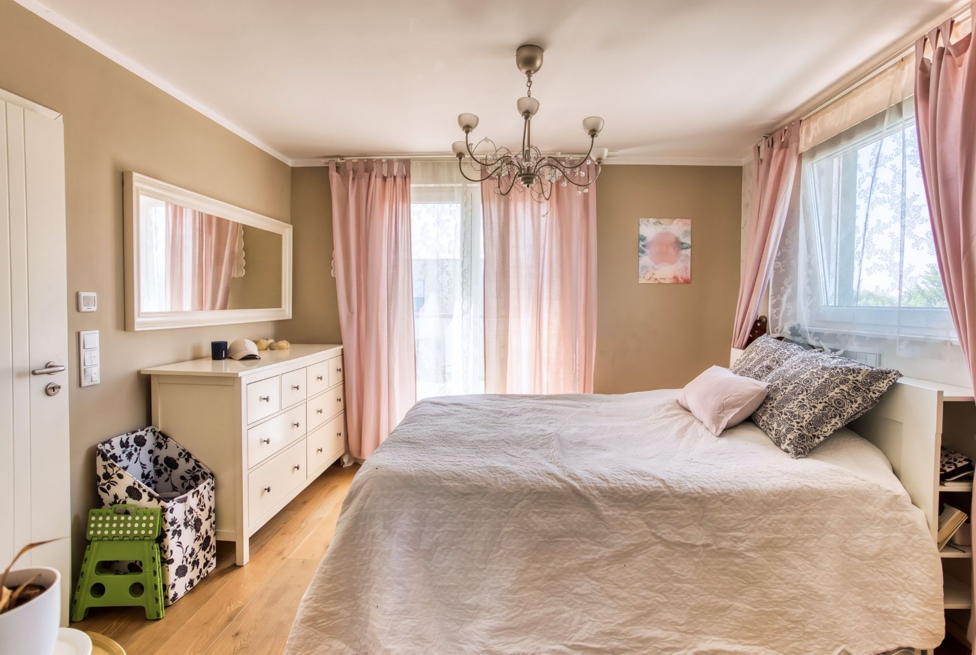 Helles Schlafzimmer mit Doppelbett, Kommode, Spiegel und rosa Vorhängen vor Fenstern.