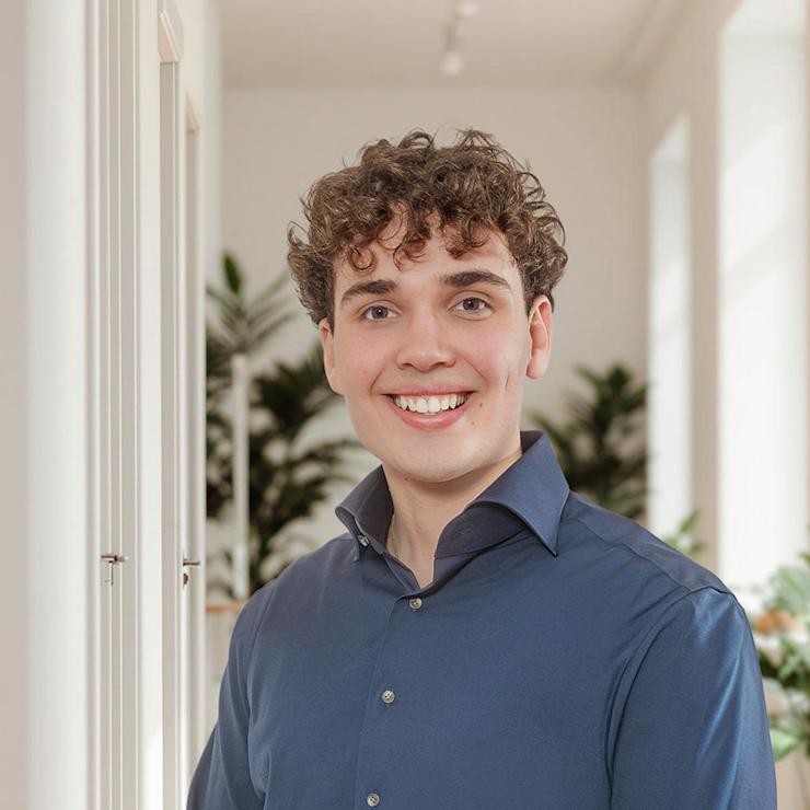 Junger Mann mit lockigem Haar steht lächelnd in einem hellen Raum mit Pflanzen im Hintergrund.
