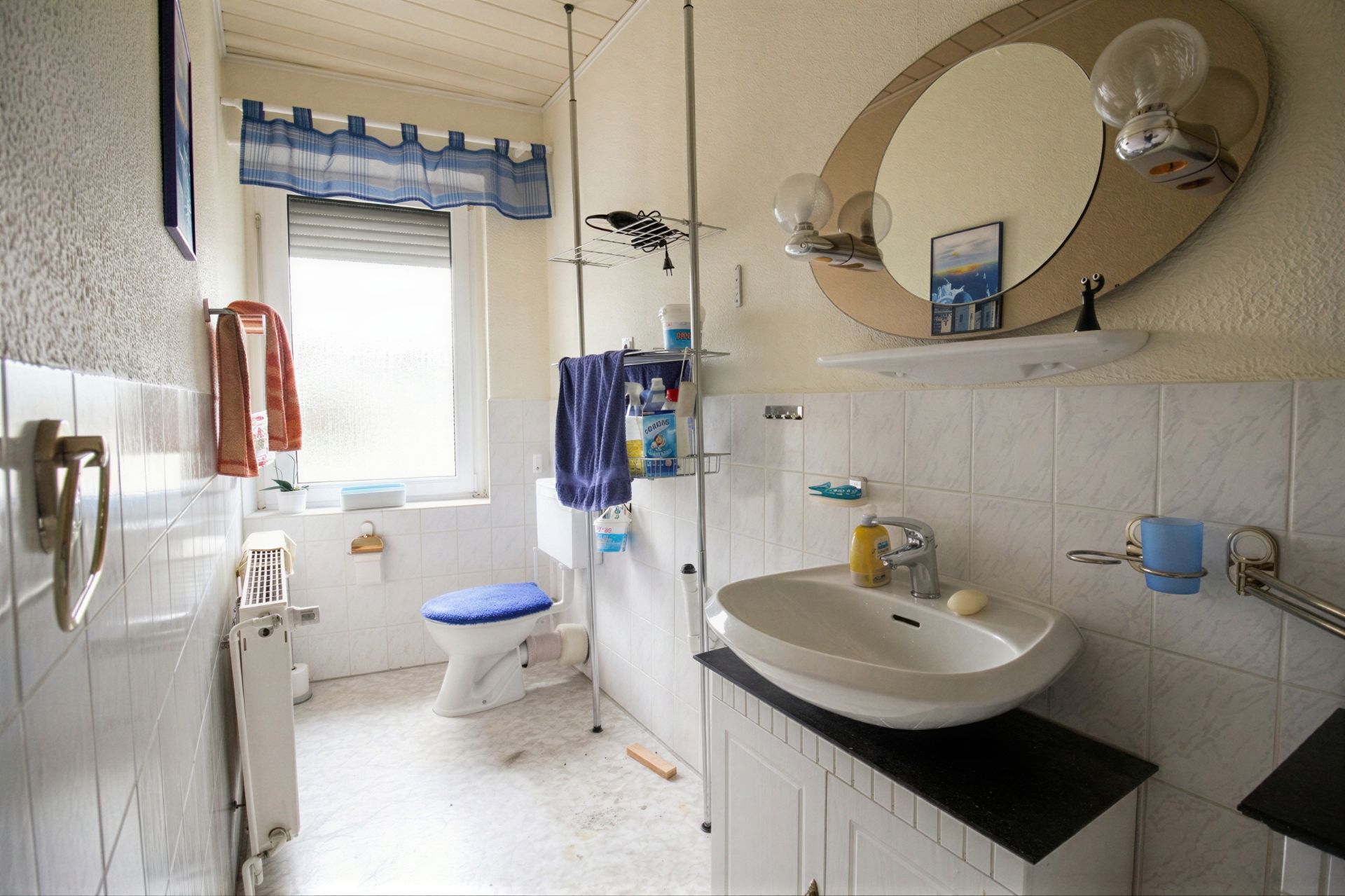 Badezimmer mit Waschbecken, Spiegel, Toilette und Fenster, Handtücher und Regale sichtbar.