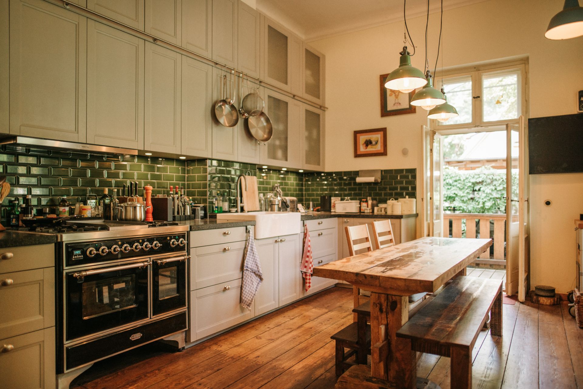 Helle Wohnküche mit Holztisch, Bänken, Herd, grüner Fliesenrückwand und offenem Zugang zum Garten.