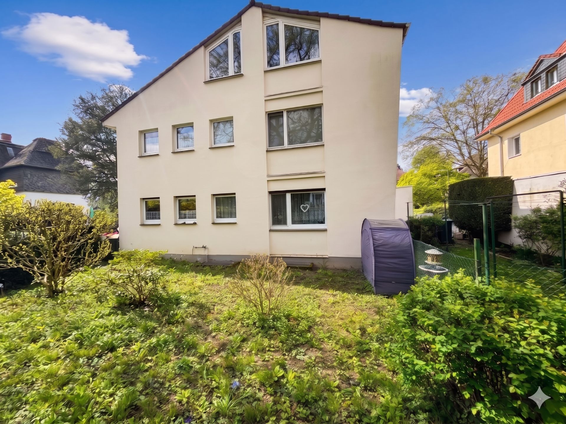 Wohnhaus mit drei Etagen und mehreren Fenstern, davor ein Garten mit Büschen.