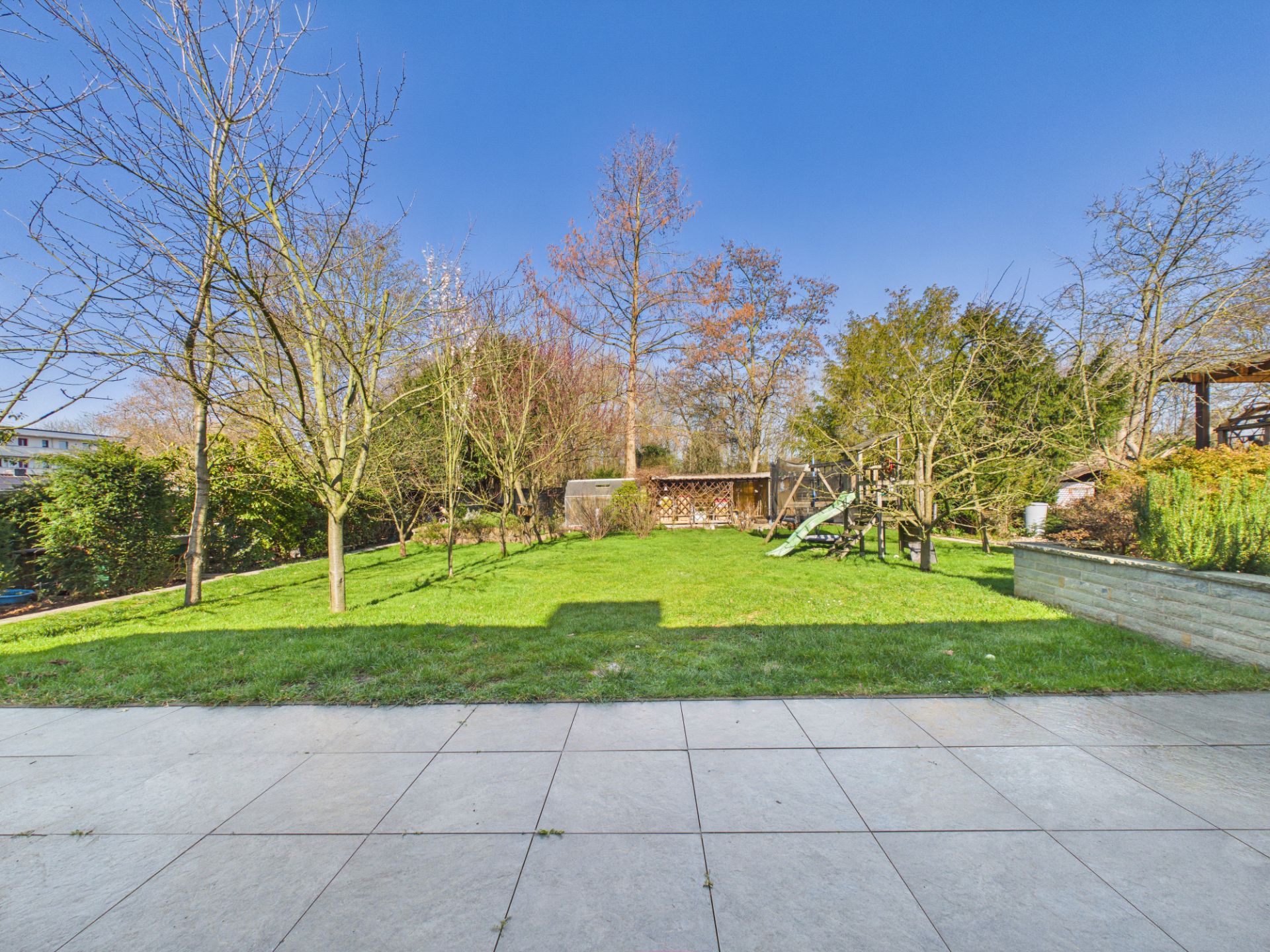 Blick auf einen Garten mit Rasen, Bäumen, Spielgerät und Terrasse im Vordergrund.