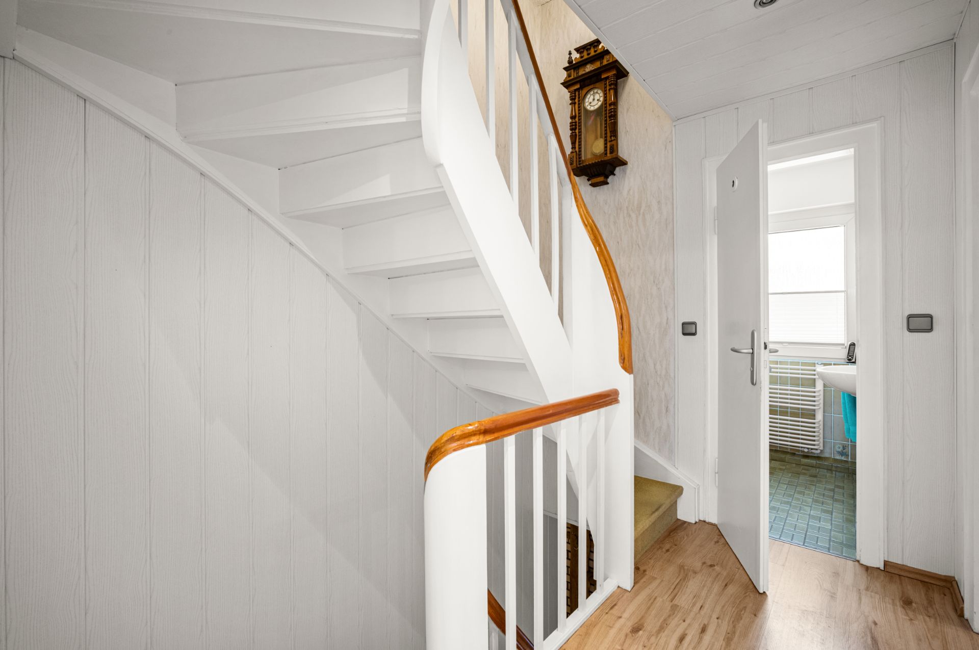 Heller Flur mit weißer Treppe, Holzhandlauf und einer Wanduhr, Tür zum Badezimmer offen.
