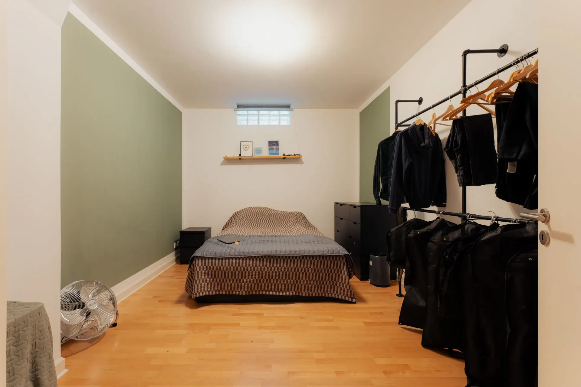 Schlafzimmer mit Bett, Kleiderstange, Kommode und Ventilator auf Holzfußboden.