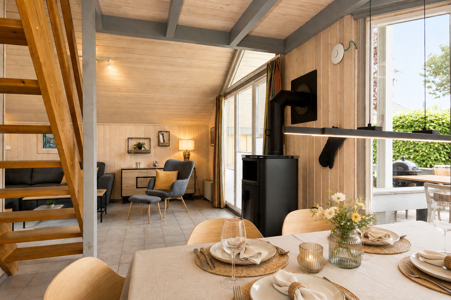 Gemütliches Wohnzimmer mit Holzwänden, Esstisch, Kamin, Treppe und großen Fenstern zur Terrasse.