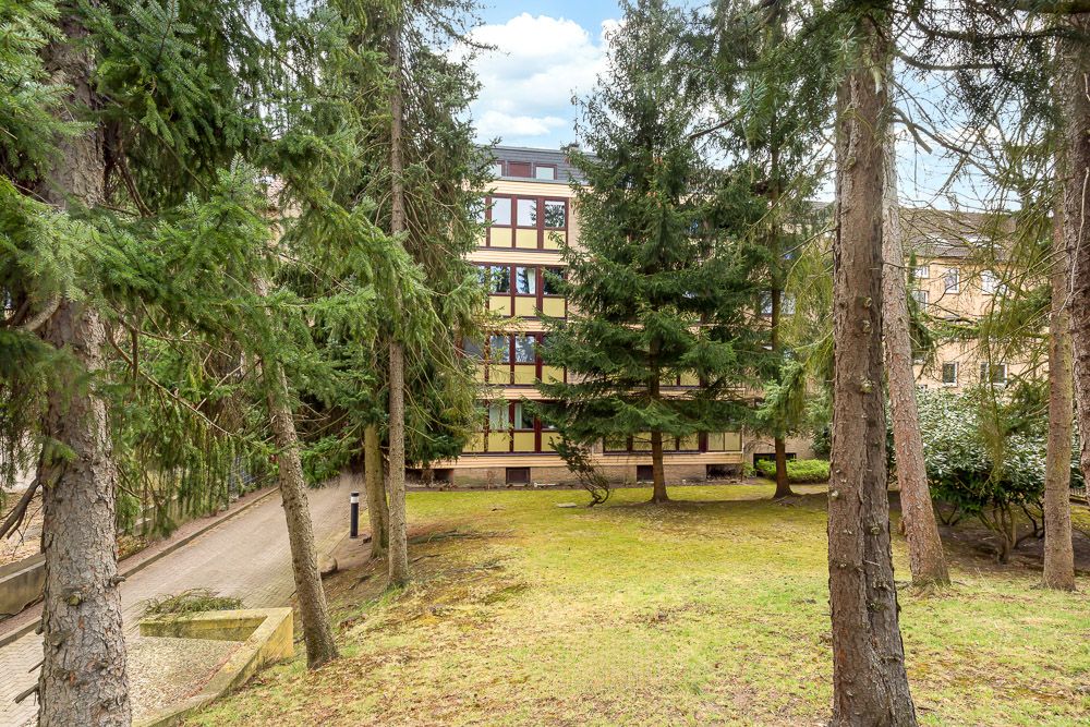 Mehrstöckiges Wohngebäude mit großen Fenstern, umgeben von Rasen und hohen Nadelbäumen.