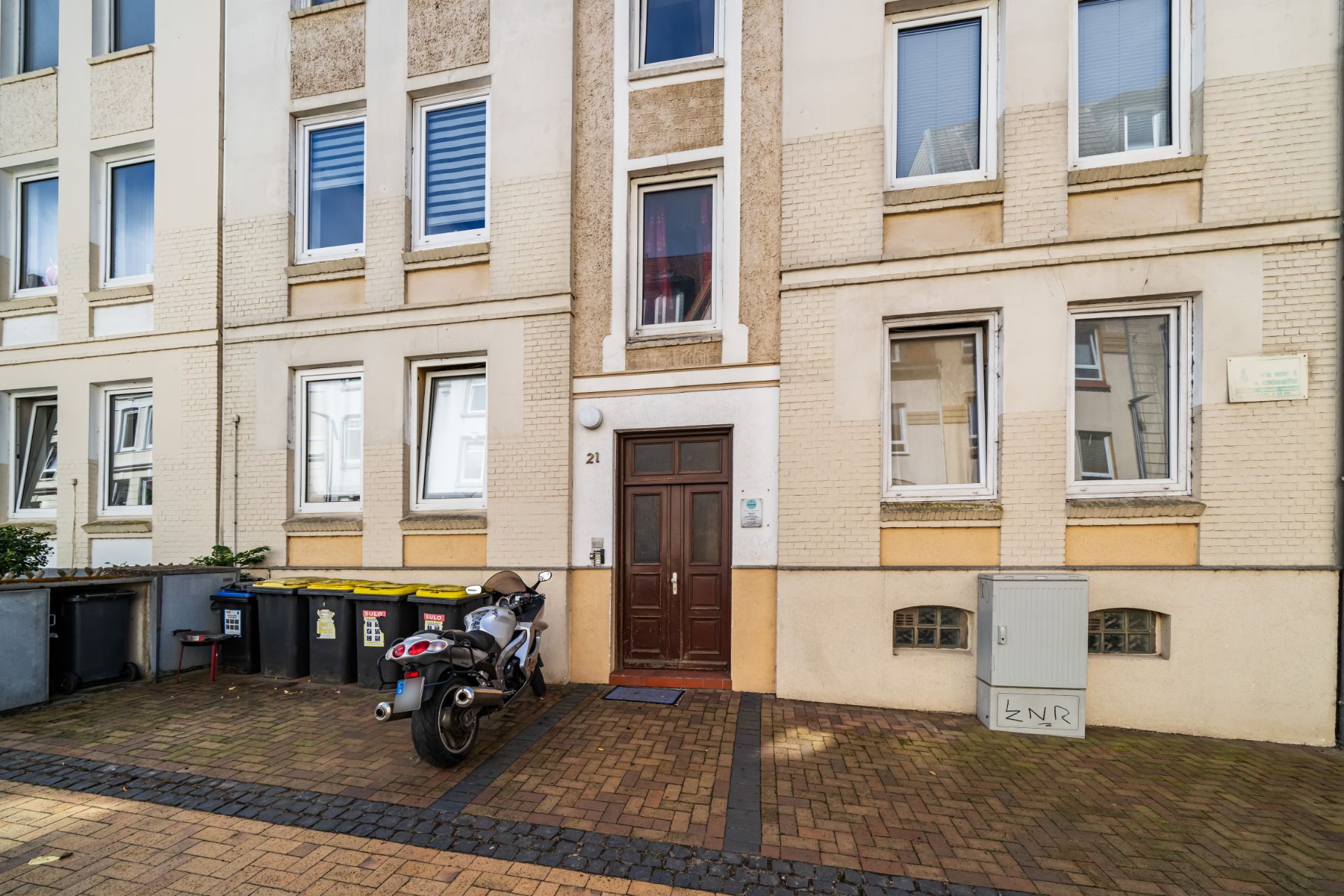 Vorderseite eines Wohngebäudes mit brauner Tür, Fenstern und geparktem Motorrad.