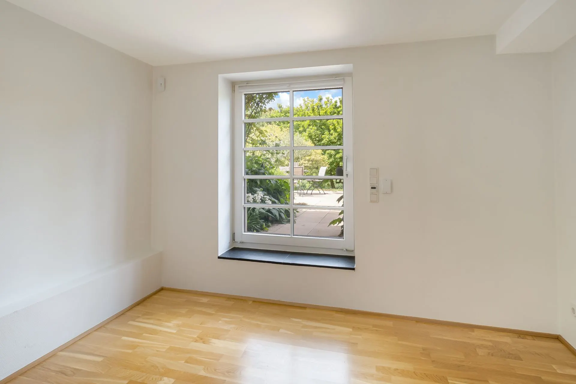 Heller Raum mit Fenster, weißer Wand und Parkettboden.