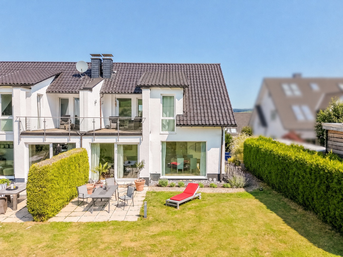 Modernes weißes Haus mit Balkon, Terrasse, Gartenmöbeln und Liege auf Rasen.