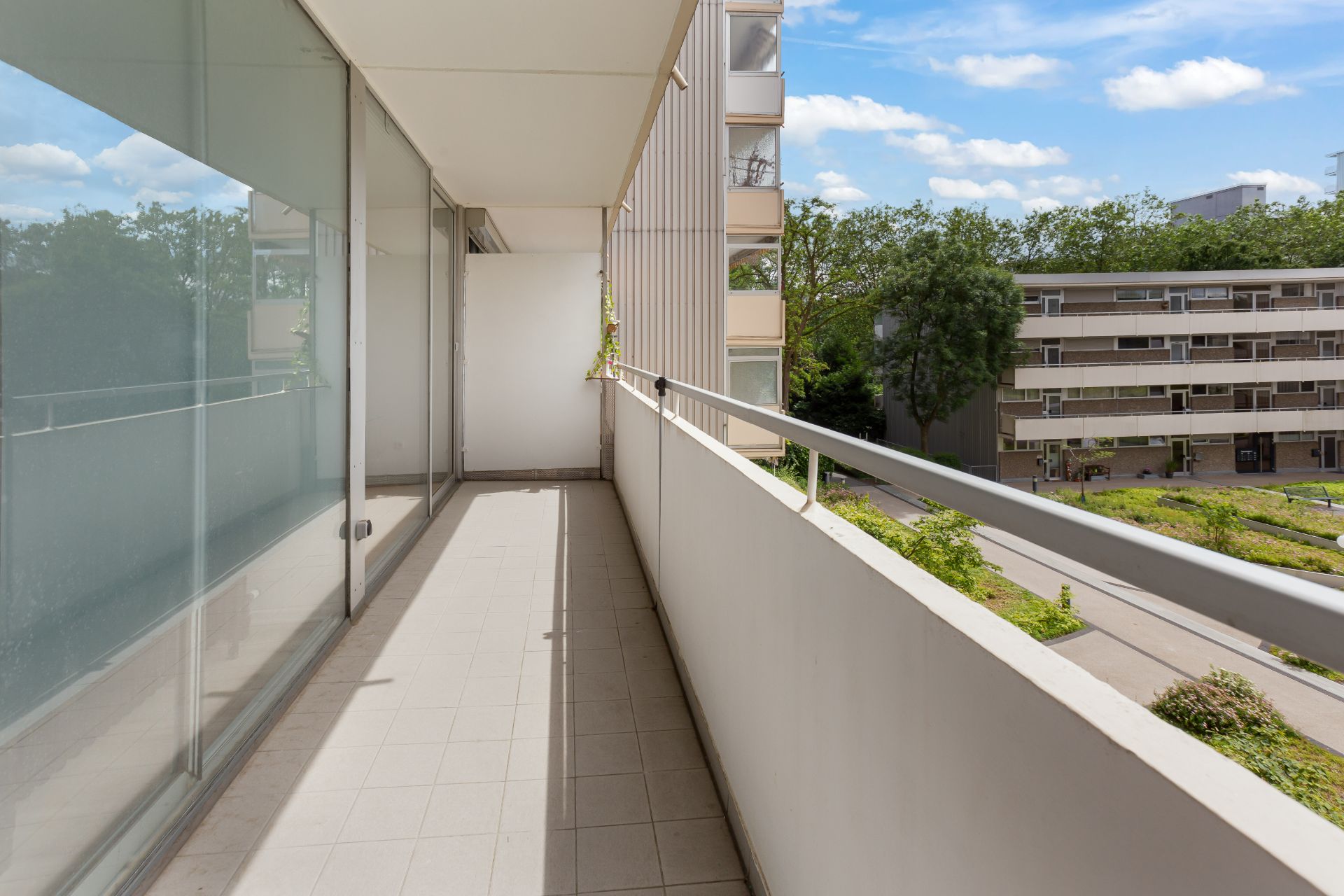 Geräumiger Balkon mit Fliesenboden und Glasgeländer, Blick auf Wohngebäude und Grünflächen.