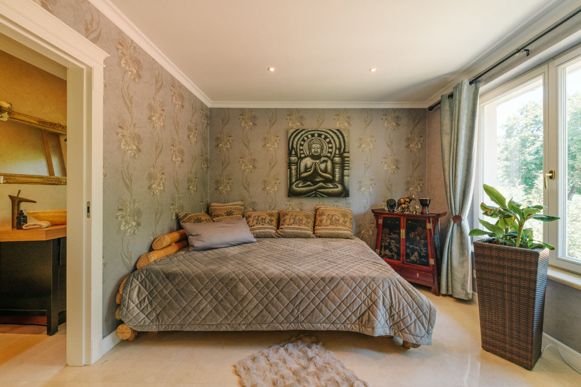 Gemütliches Schlafzimmer mit grauer Tagesdecke, Wandbild, großen Fenstern und vielen Kissen.