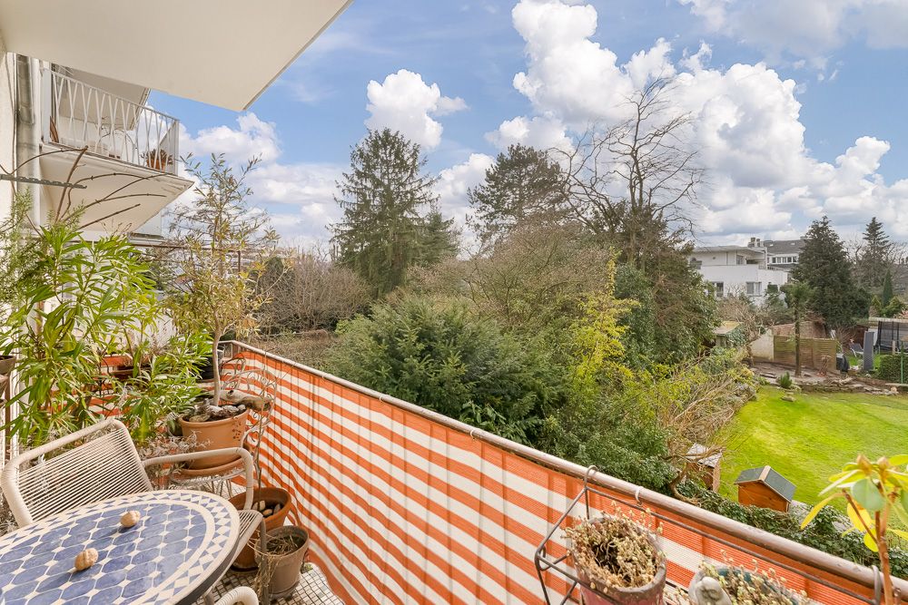 Balkon mit Pflanzen und Tisch, Blick auf grünen Garten und Bäume unter blauem Himmel.