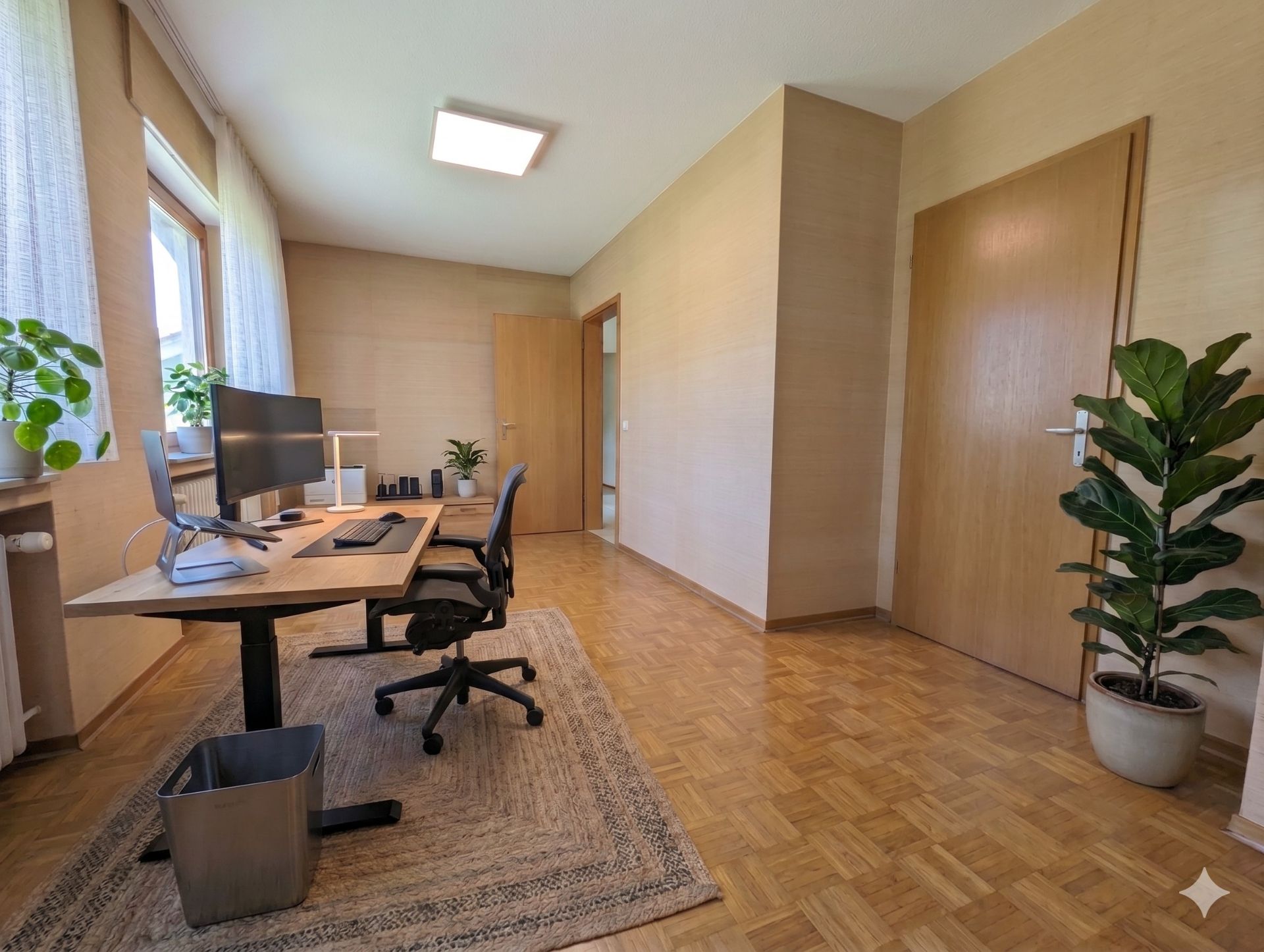 Helles Arbeitszimmer mit Schreibtisch, Stuhl, zwei Monitoren und mehreren Zimmerpflanzen.